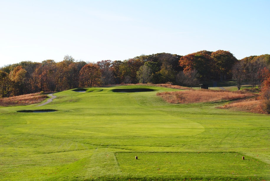 Tee box looking out into a long, wide fairway leading to bunkers and green with a border of trees just beyond.