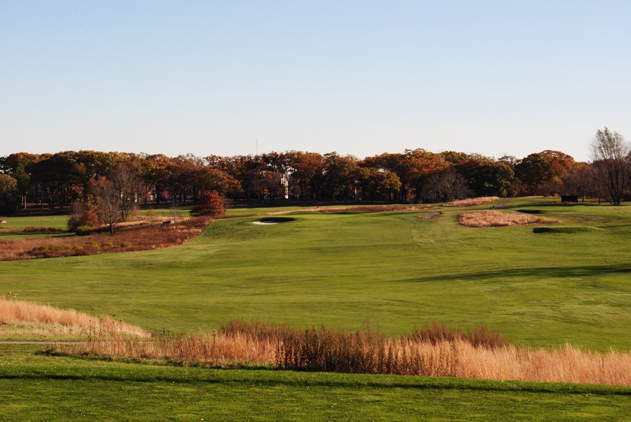 Tall grass borders the fairway in the foreground with dense woods in the background